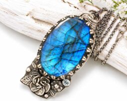 Großer ovaler Labradorit-Silberanhänger mit detailreicher Silberrose und Aquamarin neben einer geschwungenen Kette auf hellem Holz.