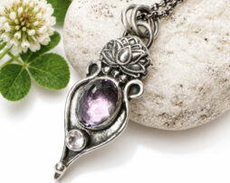Vertikale Ansicht des handgefertigten Lotus-Silberanhängers mit ovalem Amethyst und Rosenquarz auf einem hellen Stein, dekoriert mit Kleeblüten auf der linken Seite.
