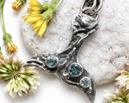 Silberner Walflossen-Anhänger mit hellblauen Aquamarin- und Topas-Steinen, liegend platziert auf einem weißen Kieselstein neben gelben Wildblumen.