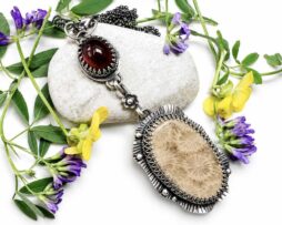 Handgefertigter silberner Statement-Anhänger mit versteinerter fossiler Koralle und rotem Granat, liegend auf einem hellen Kieselstein, dekoriert mit grünen Blättern und lila-gelben Wildblumen.