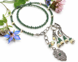 Handgemachtes Schmuckset aus 925 Sterling Silber bestehend aus einer grünen Tsavorit- und Smaragd-Halskette sowie passenden langen Ohrringen mit edlen Welo Opalen, dekorativ arrangiert neben einer blauen Blüte.