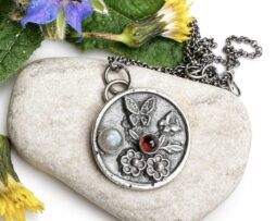 Handgefertigter runder Schmetterling-Anhänger aus 925er Silber mit Labradorit und Granat, dekoriert auf einem hellen Naturstein, umgeben von gelbem Löwenzahn und blauen Vergissmeinnicht-Blüten.