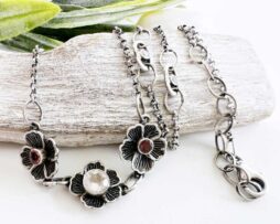 Handgefertigte Blumenkette aus Silber kombiniert romantisches Design mit hochwertigen Edelsteinen. Im Mittelpunkt des Schmuckstücks stehen drei filigran gestaltete Blüten. Die zentrale Blüte ist mit einem strahlenden Rosenquarz besetzt, während die beiden äußeren Blüten durch zarte, rosafarbene Turmaline veredelt werden. Ergänzt wird die Halskette durch fein schimmernde Moissanite, die der Kette eine besondere Raffinesse und funkelnde Akzente verleihen. Der Rosenquarz symbolisiert Liebe, Harmonie und Wärme des Herzens, während dem Turmalin nachgesagt wird, Lebensfreude und positive Energie zu fördern. Die funkelnden Moissanite setzen durch ihr beeindruckendes Lichtspiel elegante Glanzpunkte.