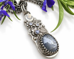 Schräge Makroaufnahme des floralen Silber-Anhängers mit funkelndem Sternsaphir vor echten blauen Blüten.